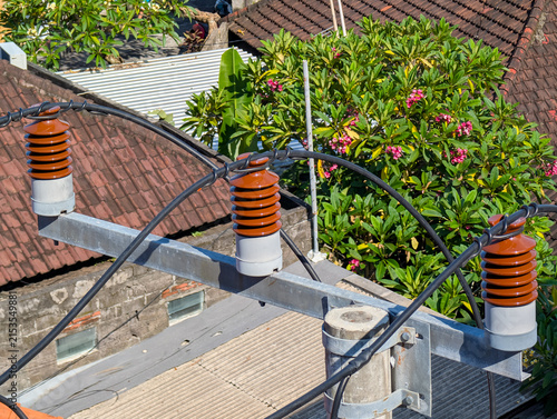 Foto High-voltage ceramic insulators mounted on a utility pole crossarm supporting power lines above rooftops and green trees