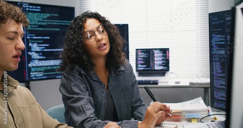 Two Hispanic software engineers pair-program AI models at a shared workstation, reviewing datasets and model metrics on multiple monitors. They discuss optimization strategies, version control, and de