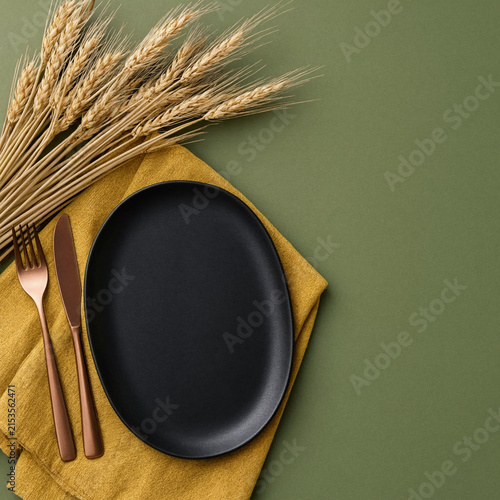 Top view of empty black plate with cutlery and wheat on green background