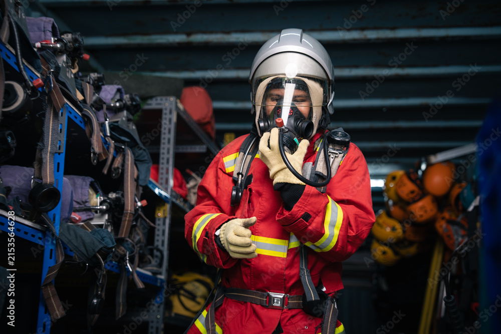 Fototapeta premium Firefighter in Full Protective Uniform Giving a Thumbs-Up Gesture while Adjusting a Face Mask inside an Equipment Storage Area