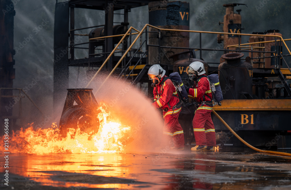 Naklejka premium Firefighter Team Battling an Industrial Fire with a Hose while Wearing Full Protective Uniforms, Face Masks, Helmets, and Air Tanks