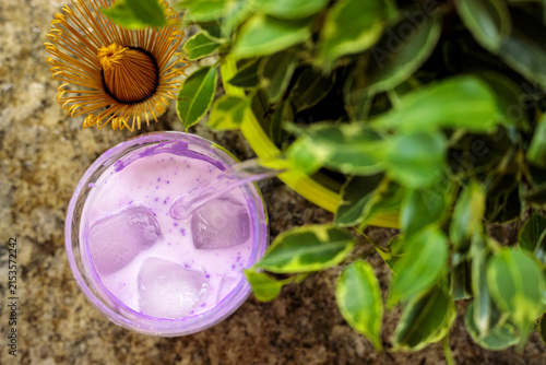Top view of a pink ube drink with ice in a glass, styled with a metal strainer, dried flower, and greenery on a stone surface