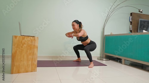 Mexican woman doing squats at home following an online fitness class on her laptop in the living room for a healthy lifestyle