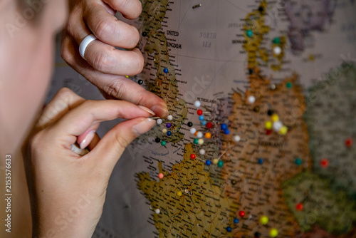 Close-up of a person's hand placing a colorful pushpin into a wall map of Europe and the United Kingdom, representing travel planning and tracking destinations. The map prominently shows Europe