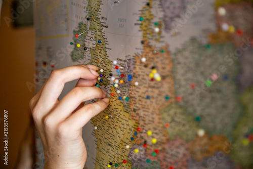 Close-up of a person's hand placing a colorful pushpin into a wall map of Europe and the United Kingdom, representing travel planning and tracking destinations. The map prominently shows Europe