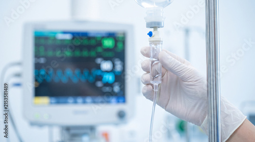 Gloved hand adjusting IV drip in hospital with patient monitor in background, showing critical care concept