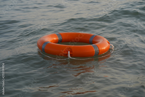 Orange life ring floats gently on calm water in ocean. Ripples surround the buoy, suggesting gentle movement. Its stripes and knot reflect safety and rescue