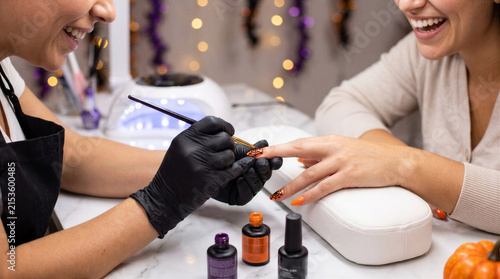 Woman receiving Halloween themed manicure from nail technician in decorated salon. Seasonal beauty service with orange spiderweb nail art, festive retail and self care concept