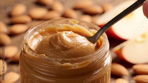 Close up of a spoon dipping into creamy peanut butter jar with almonds and apple
