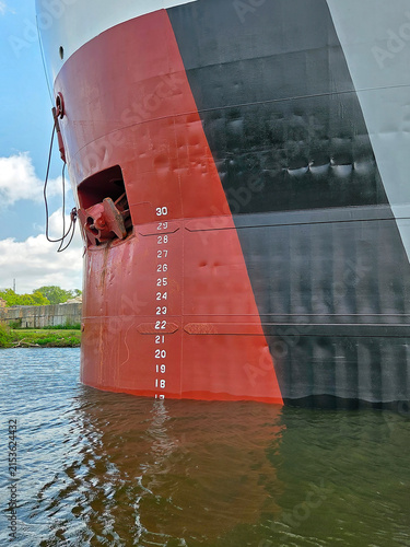 A close-up view of a Great Lakes freighter's bow features a distinctive red and black color scheme, with visible plimsoll markers indicating the highest safe depth for the ship in the water