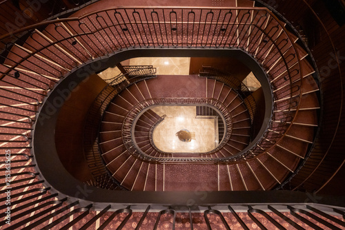 View of the elegant elliptical spiral staircase with dark red carpeting and ornate railings inside the Tokyo Kaikan Main Building in Chiyoda City, Tokyo, Japan.