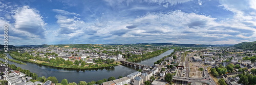 Luftbild von Trier mit der antiken Römerbrücke über die Mosel, umgeben von Stadtarchitektur und grünen Hügeln unter bewölktem Himmel an einem Sommertag.