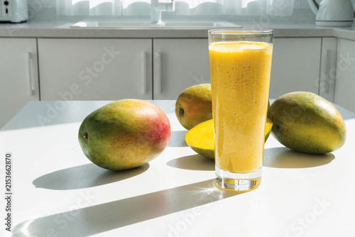 Fresh yellow mangos and glass of mango smoothie on the white table in the white modern kitchen. Copy space