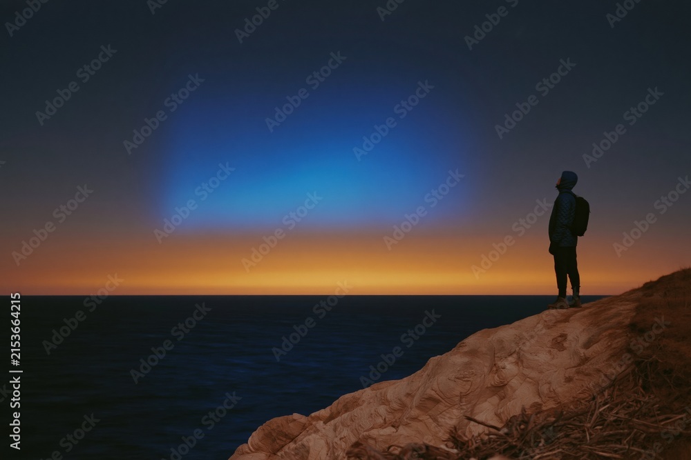 Fototapeta premium Lone person in a hooded jacket standing on a weathered stone cliff looking at a glowing blue light in the night sky for travel and adventure promotion
