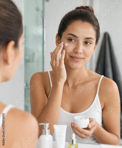 Young woman applying moisturizer cream on her face while looking in the bathroom mirror during morning routine