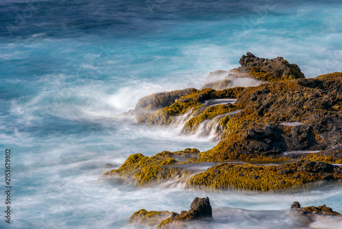 Long exposure of ocean waves crashing on volcanic rocks covered with green algae