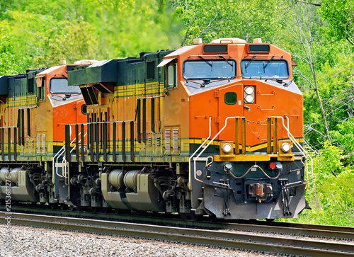 Two orange and black diesel‑electric train locomotives traveling through a green wooded corridor on a rural railroad line