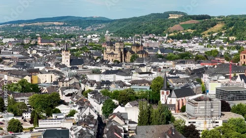 Luftbild von Trier mit der antiken Römerbrücke über die Mosel, umgeben von Stadtarchitektur und grünen Hügeln unter bewölktem Himmel an einem Sommertag.