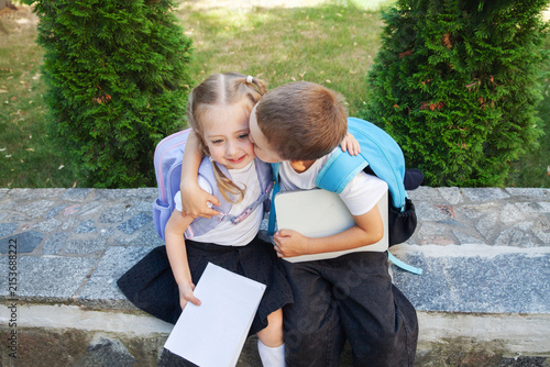 Brother  kisses his sister on the cheek as they sit outdoors with backpacks and a digital tablet, celebrating their back to school journey