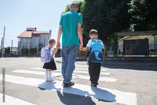 Father walks his son and daughter across a crosswalk on the way to school. Children with backpacks head to class on a sunny morning
