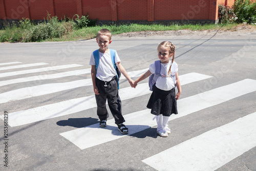 Brother and sister holding hands safely cross a pedestrian crossing with backpacks on their way to school on a sunny morning