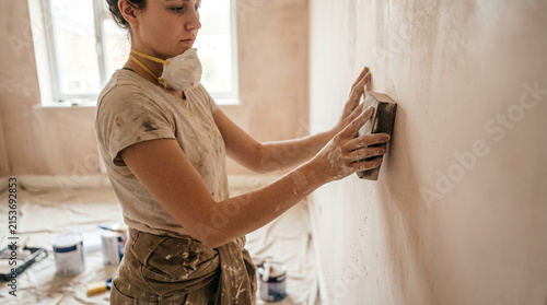 Female decorator sanding plastered wall during home improvement project in an empty room.