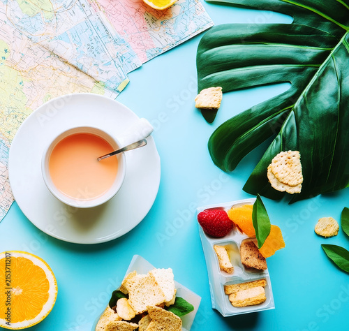 A refreshing snack setup featuring a cup of light orange liquid, fresh orange slices, and dried fruits beside a verdant monstera leaf, ideal for a tropical, laid-back breakfast