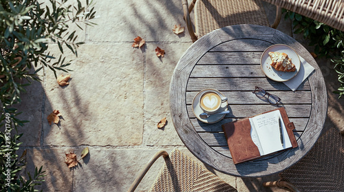 Top view of coffee and pastry on wooden outdoor table with book and olive leaves, cozy mediterranean patio breakfast morning scene