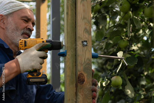 A man uses a cordless screwdriver to attach a wooden beam to a metal frame. He uses a clamping tool called a tie rod to temporarily secure the beam.