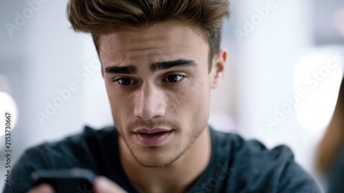 Young man with thoughtful expression looking intently at smartphone screen while processing important information indoors in soft natural light