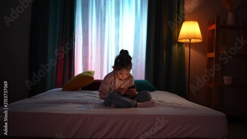 A child sitting on a bed in a dark room in the evening, using a smartphone.