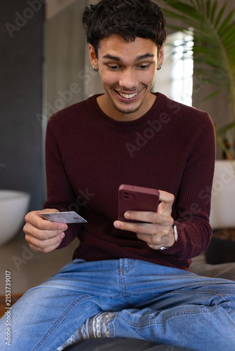 African American man in 20s wearing burgundy sweater holding smartphone and card on cushion at home