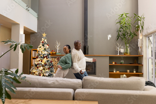African American couple standing back-to-back in living room with lit Christmas tree, fireplace