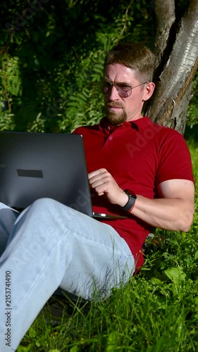 Busy man working on a laptop while sitting on the grass in a city park sitting under tree.