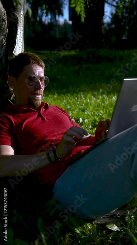 Busy man working on a laptop while sitting on the grass in a city park sitting under tree.