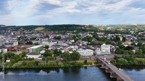 Zeitraffer (Timelapse)  Drohnenaufnahme von Trier mit der antiken Römerbrücke über die Mosel, umgeben von Stadtarchitektur und grünen Hügeln unter bewölktem Himmel an einem Sommertag.
