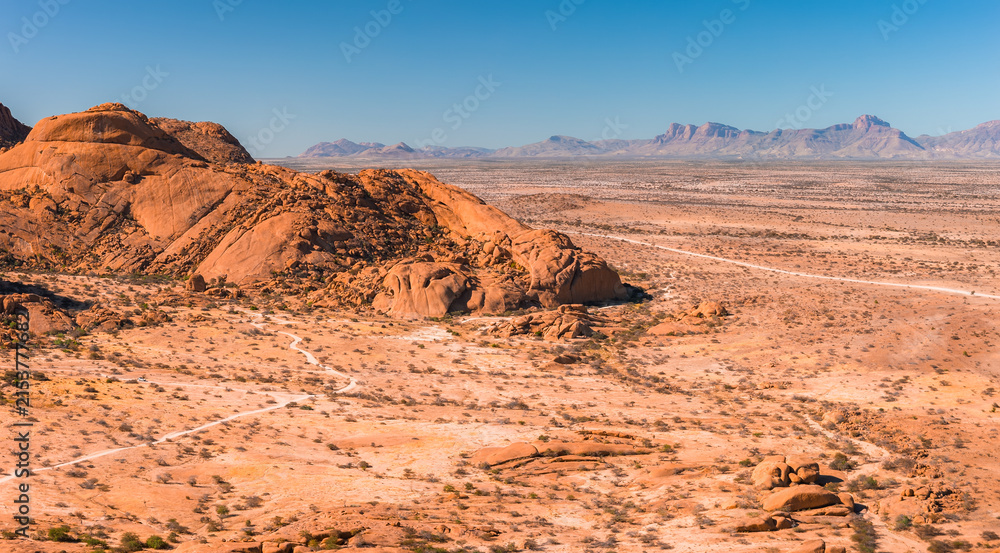 Obraz premium An aerial perspective shows the vast desert landscape of Spitzkoppe in Namibia, featuring large granite rock formations, dry plains, and distant mountains.