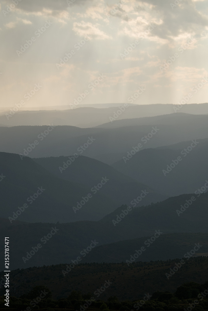 Obraz premium Dramatic mountain landscape with sun rays shining through storm clouds and distant rain over layered hills.