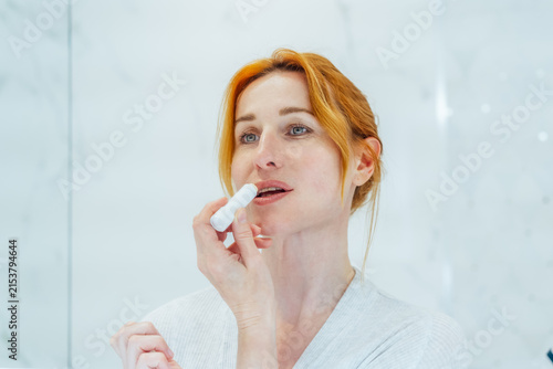 Middle aged woman applies lip balm looking in mirror in white bathroom. SPF Lip Balm for lips kin protection. Home beauty treatment and skin care routine.