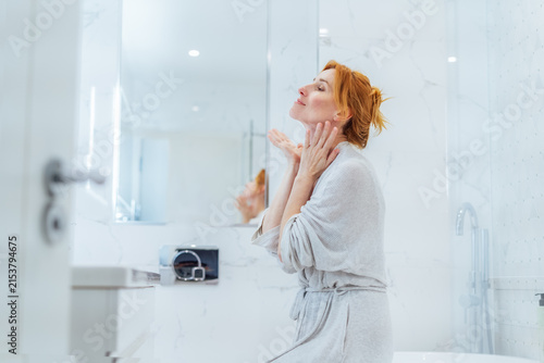Middle aged woman doing face building yoga facial gymnastics self massage and rejuvenating exercises looking in bathroom mirror. Natural procedures for skin care. Home Beauty treatment routine