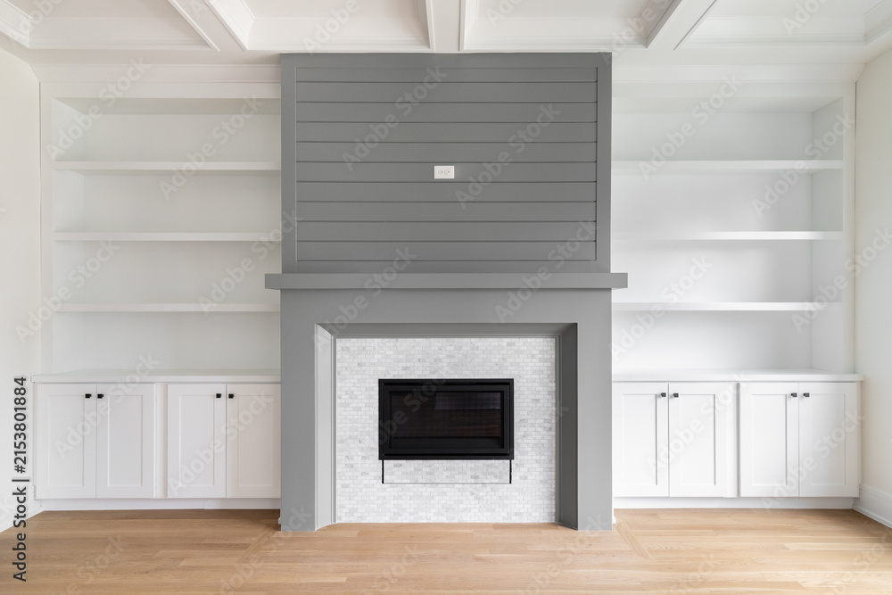 Fototapeta premium A fireplace detail with white built-in shelving and cabinets, white oak flooring, white coffered ceiling, and a grey accent surround and mantel.
