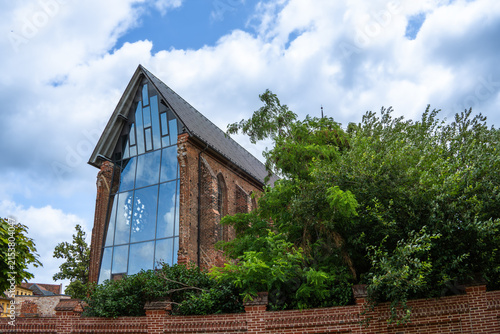 Historic brick church with a modern glass facade, framed by trees and a brick wall under a cloudy summer sky
