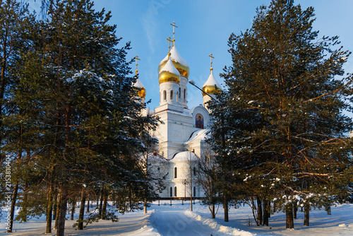Fotografie White orthodox church with golden domes surrounded by snowy pine trees on a sunny winter day