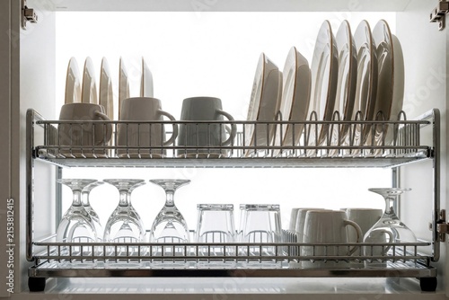 Organized kitchen rack holds clean plates, cups, and glasses on chrome shelves. Neatly stacked dishes and drinkware reflect bright light, showing tidy dishware presentation.