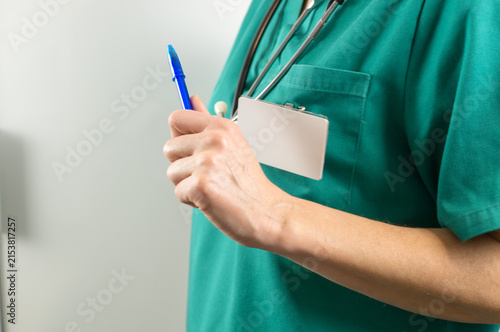 Close-up image of a nurse or doctor holding a blue ballpoint pen, dressed in green medical scrubs with a blank card badge.