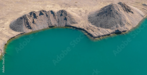 Aerial view of a turquoise water-filled mining excavation surrounded by steep piles of extracted rock and barren terrain at an active mining site, Le Pouzin, Auvergne-Rhone-Alpes, France.
