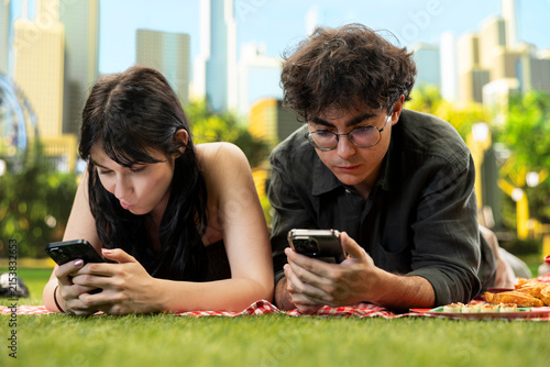Fotografie Young couple lie side by side on a picnic blanket and ignore each other while doomscrolling on smartphones, showing phubbing, poor communication, digital distraction, and relationship disconnection