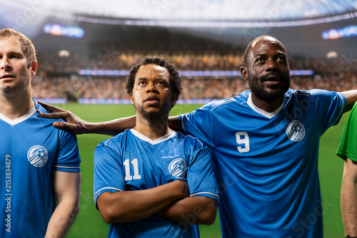 Diverse soccer players and goalkeeper stand together in a pre-match anthem lineup on the pitch, showing team unity with the stadium crowd blurred behind them during the opening ceremony.