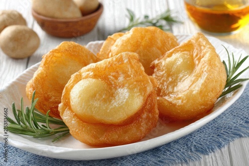 Crispy golden-brown fritters, likely potato, on a white plate, with sprigs of rosemary.  Potatoes in a bowl in the background