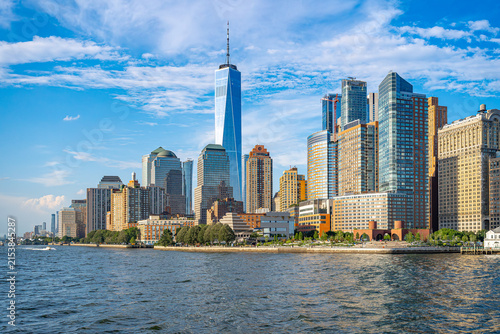 Financial district skyline along Hudson River in New York City. Tall office buildings and green waterfront park stretch under blue sky.